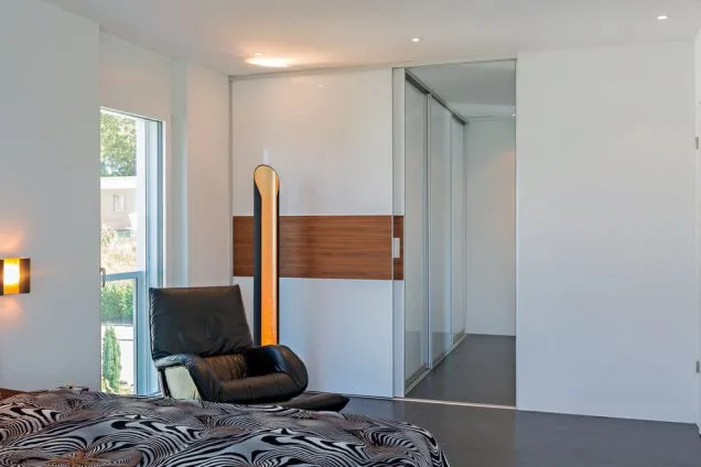 Schiebetüre als Raumtrenner für Ankleide mit Querfries in Holz 1 Ein Raumteiler, welcher nur mit seiner rechten Seite beweglich ist und den Durchgang auf einen Gang mit angrenzenden Schiebetüren freigibt. Davor befindet sich seitlich ein gemütlich wirkender Sessel. Der Raumteiler besteht aus weißen Hochglanzelementen oben und unten und in der Mitte aus einem edlen Furnier.