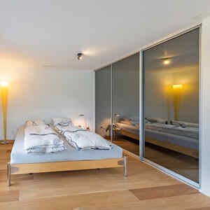 Ein Designerbett mit einem Einbauschrank neben sich. Der Einbauschrank ist komplett geschlossen und in seinen Schiebetüren spiegelt sich das Licht einer Lampe aus dem Schlafzimmer.