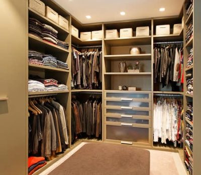 Ankleidezimmer in Besch mit edler Beleuchtung, Drehtürenkästen, dem Seitensystem kombiniert mit mehreren Regalen und Schubladen in moderner edler Optik. In der Mitte befindet sich noch eine Sitzgelegenheit.