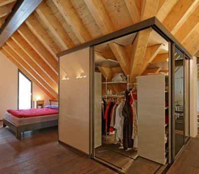 Schrank unter der Dachschräge. welcher sogar um ein Eck herum geht. Der Schrank besteht aus einer Schiebetürfront über Eck aus verspiegelten SChiebetüren und weißen Hochglanzschiebetüren. Auf diesem Bild ist der geöffnete Schrank unter der Dachschräge abgebildet, der einen Ausblick auf dessen Innenleben aus Schieberegalen und unserem Säulensystem kombiniert mit Kleiderstangen und in einer doppelten Tiefe unter der Dachschräge abgebildet.