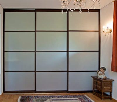 Satinierte Acrylglasschiebetüren, welche in dunklem Holz gerahmt sind teilen hier optisch zwei Räume voneinander. Die Schiebetüren sind geschlossen und erlauben keinen Blick in den nächsten Raum. Rechts vor der Schiebetür befindet sich noch ein kleines Kästchen mit einem Engel und davor befindet sich ein gemütlich wirkender Teppich.